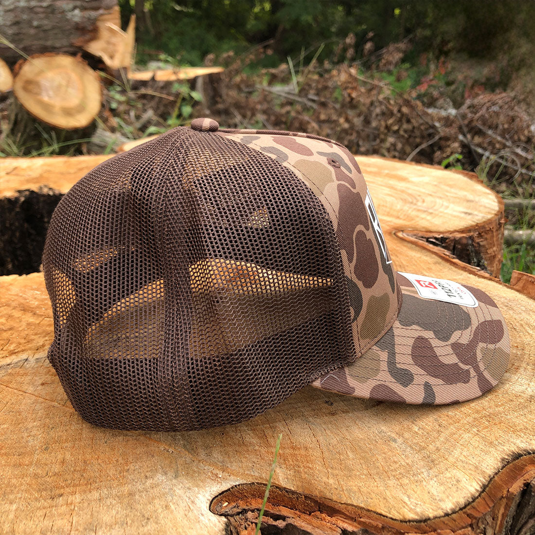 A brown and camouflage trucker hat with a mesh back, resting on a cut tree stump outdoors.