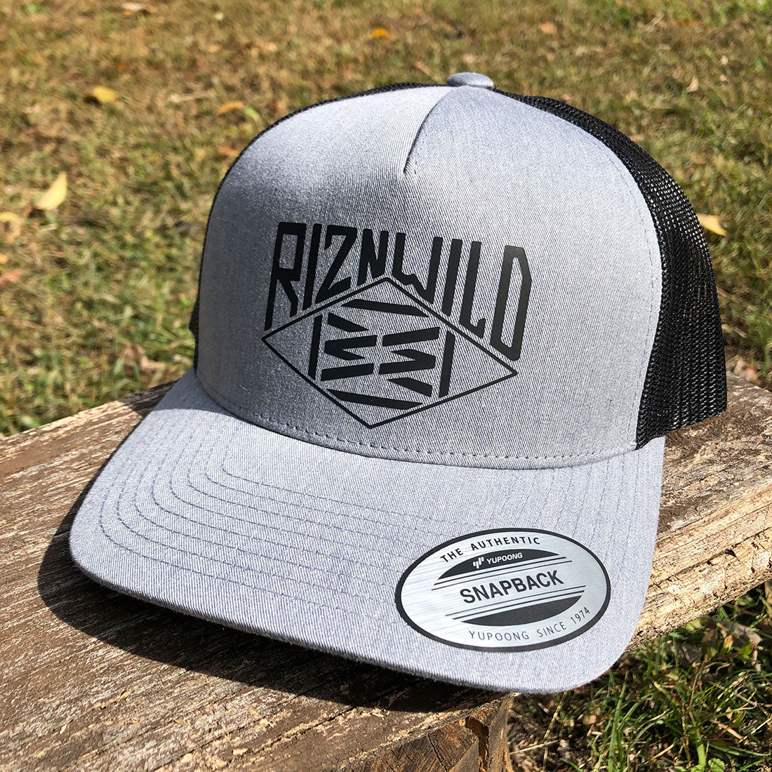 Heather grey and black RIZNWILD trucker hat with front logo, displayed outdoors on a wooden bench.