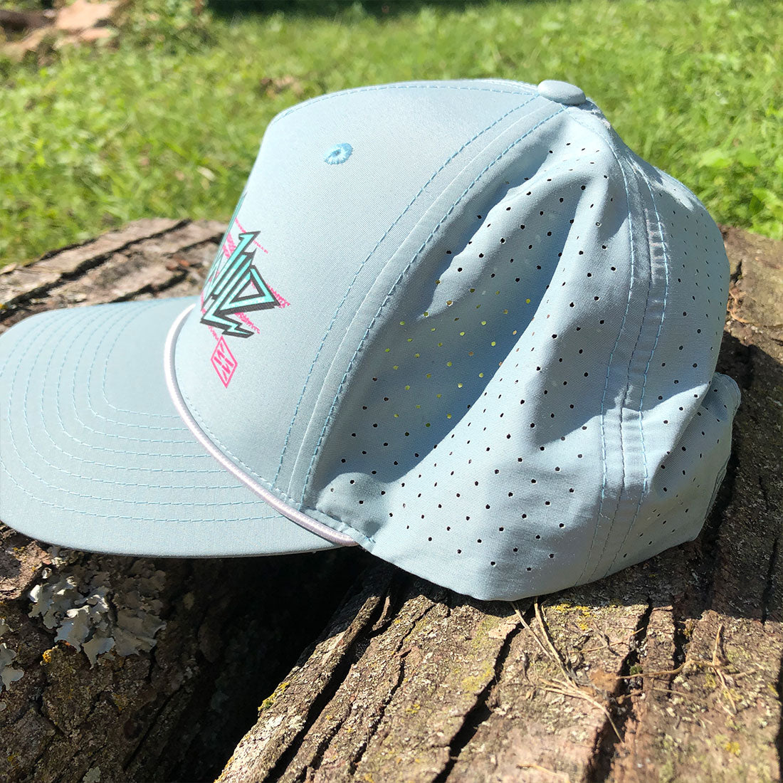 A light blue perforated baseball cap resting on a weathered tree stump outdoors, with green grass in the background. The cap features a stylized graphic logo on the front panel.