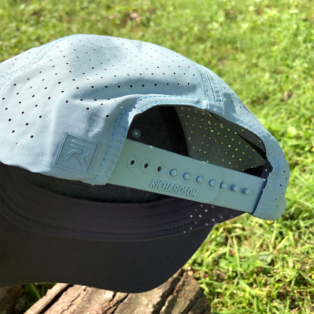 A light blue perforated baseball cap with a dark gray visor, photographed outdoors on a grassy lawn. The back strap is light blue with 'RICHARDSON' printed on it and a brand logo patch on the side.