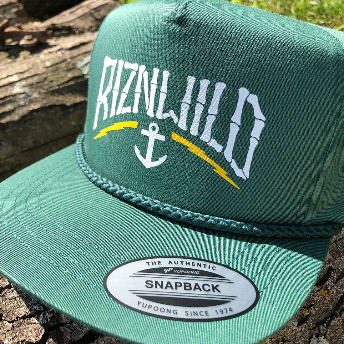 A green snapback cap with 'RIZNWILD' in white arched lettering and a white anchor graphic with yellow curved accents on the front. The brim has a braided green rope detail and a white Yupoong Snapback sticker.