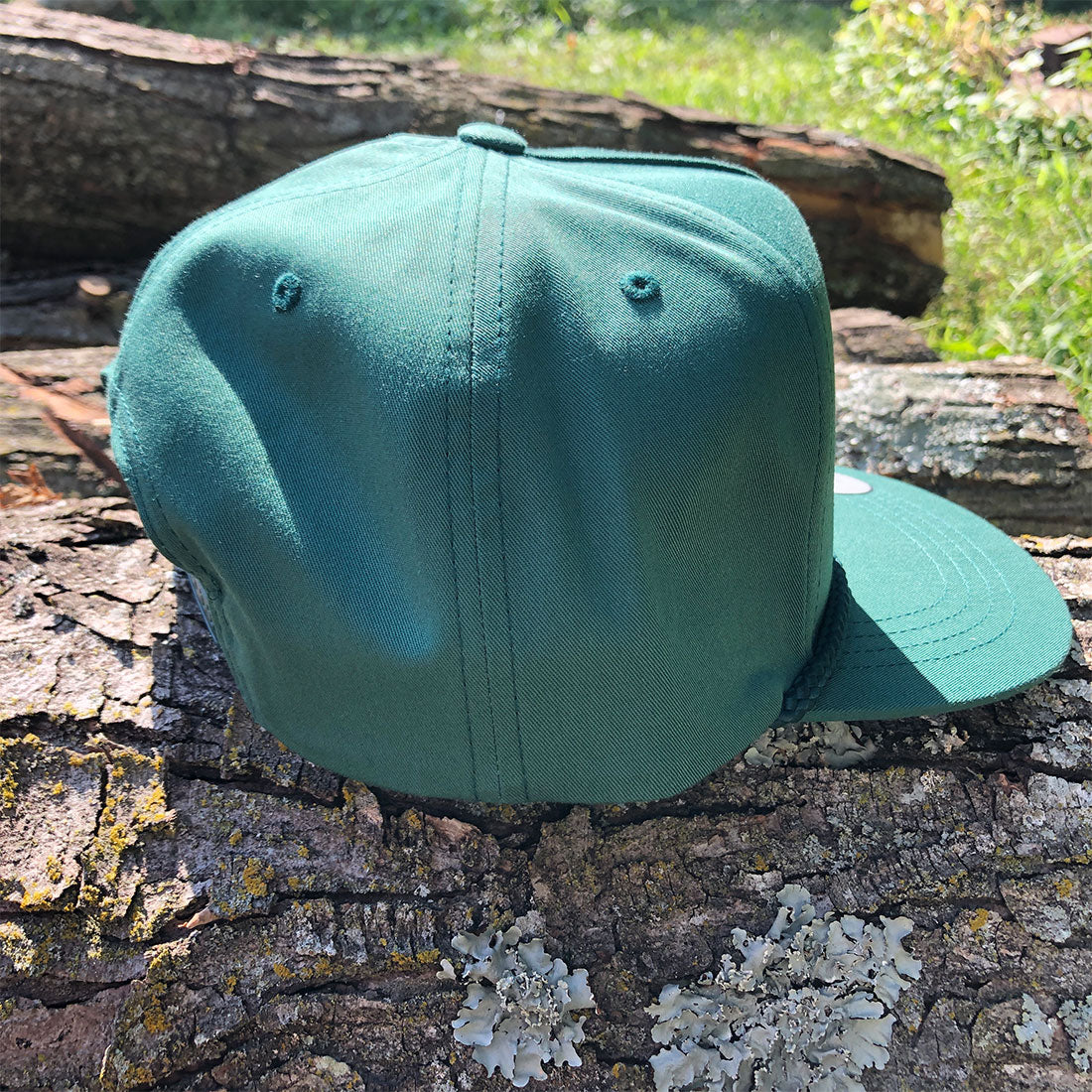 A green flat-brim baseball cap resting on a textured log outdoors with grass and logs in the background.