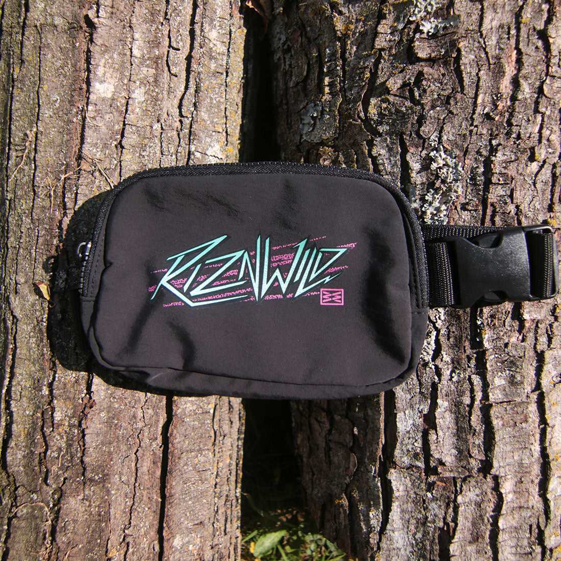 A black waist bag with a colorful 'RIZNWILD' graphic, attached to a black strap and photographed against a textured tree trunk.