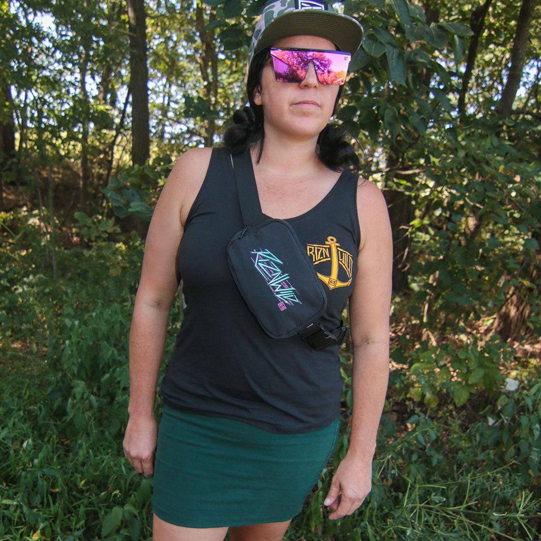 A woman wearing a black tank top, green skirt, and a black crossbody fanny pack standing outdoors in a wooded area.