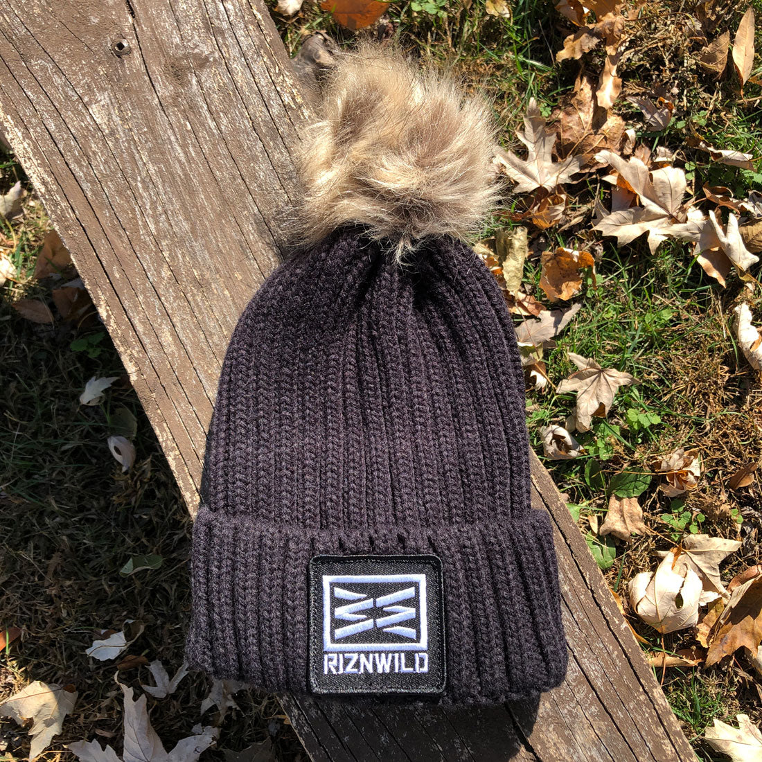 Black RIZNWILD faux fur pom beanie on a rustic wooden bench surrounded by fall leaves in sunlight