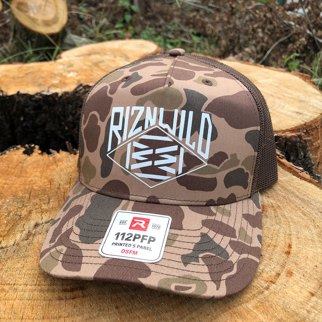 A camouflage trucker hat with a mesh back, featuring a large white 'RIZNWILD' logo on the front and a '112PFP' sticker on the brim, displayed on a cut wood log with trees in the background.
