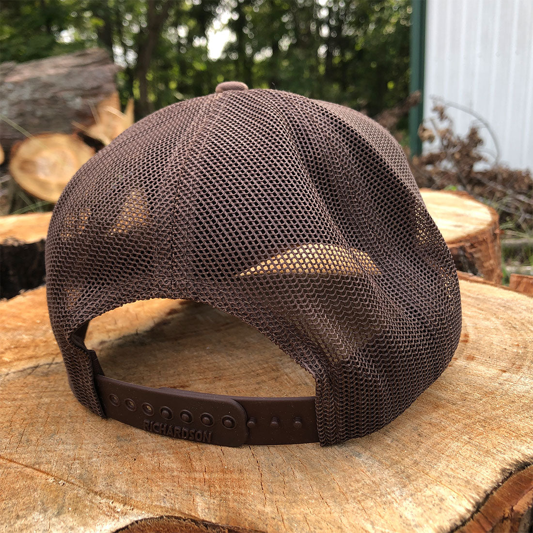 A brown mesh-back baseball cap with an adjustable brown strap, resting on a cut wood stump with a forest background.