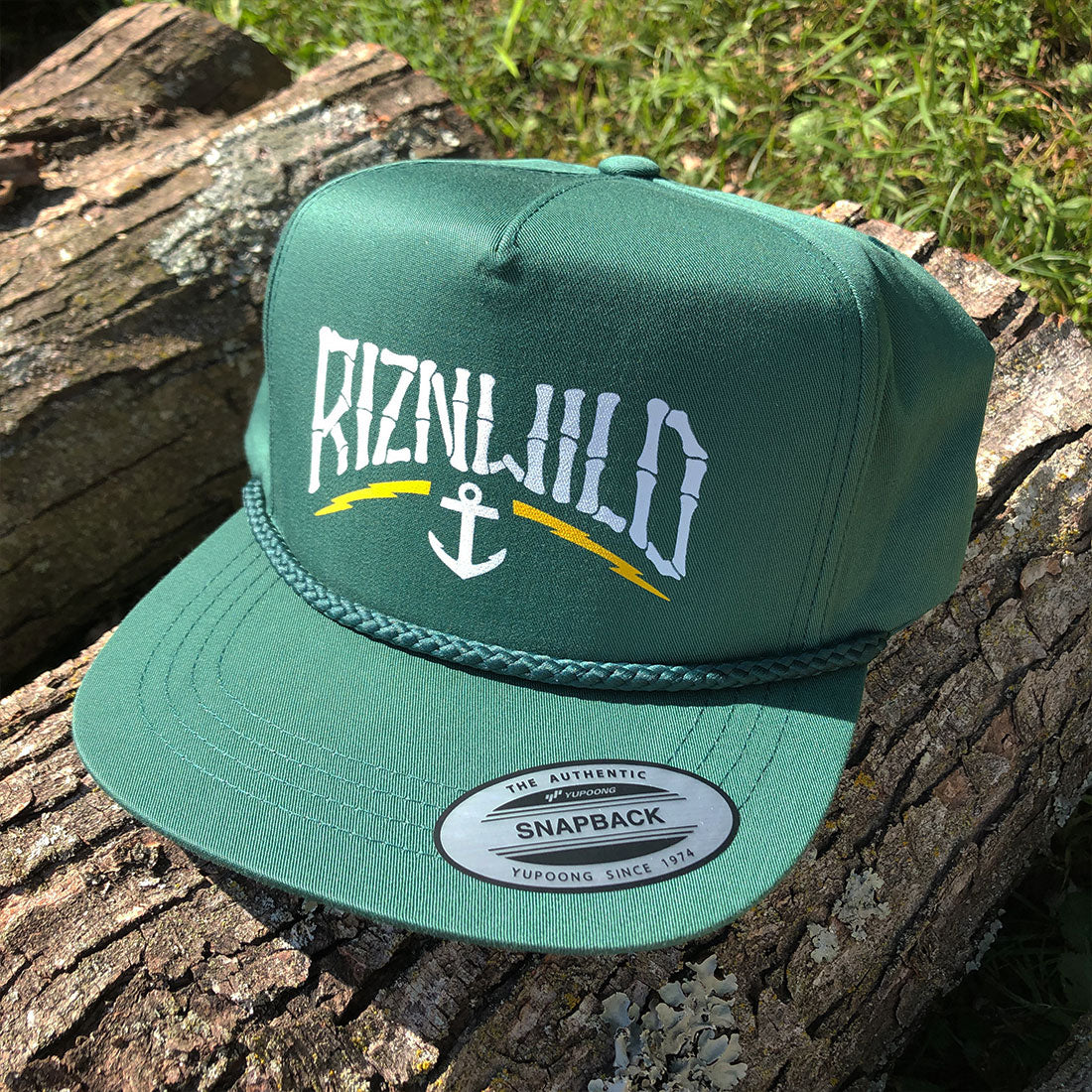 A green snapback cap with a white 'RIZNWILD' graphic and anchor logo on the front, resting on a log outdoors with grass in the background.