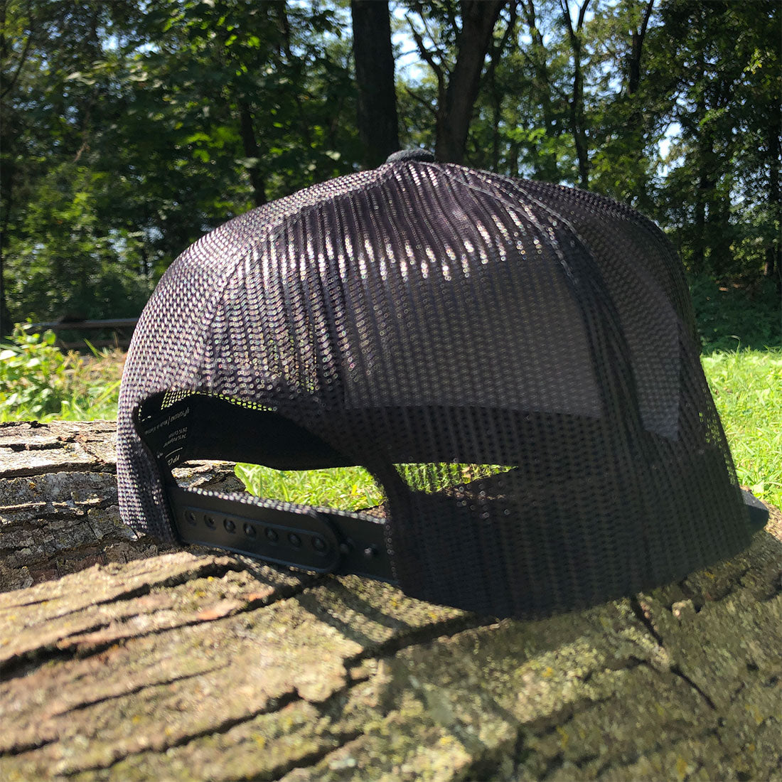 A black mesh trucker cap with an adjustable black strap, resting on a weathered tree stump outdoors with a background of green grass and trees.