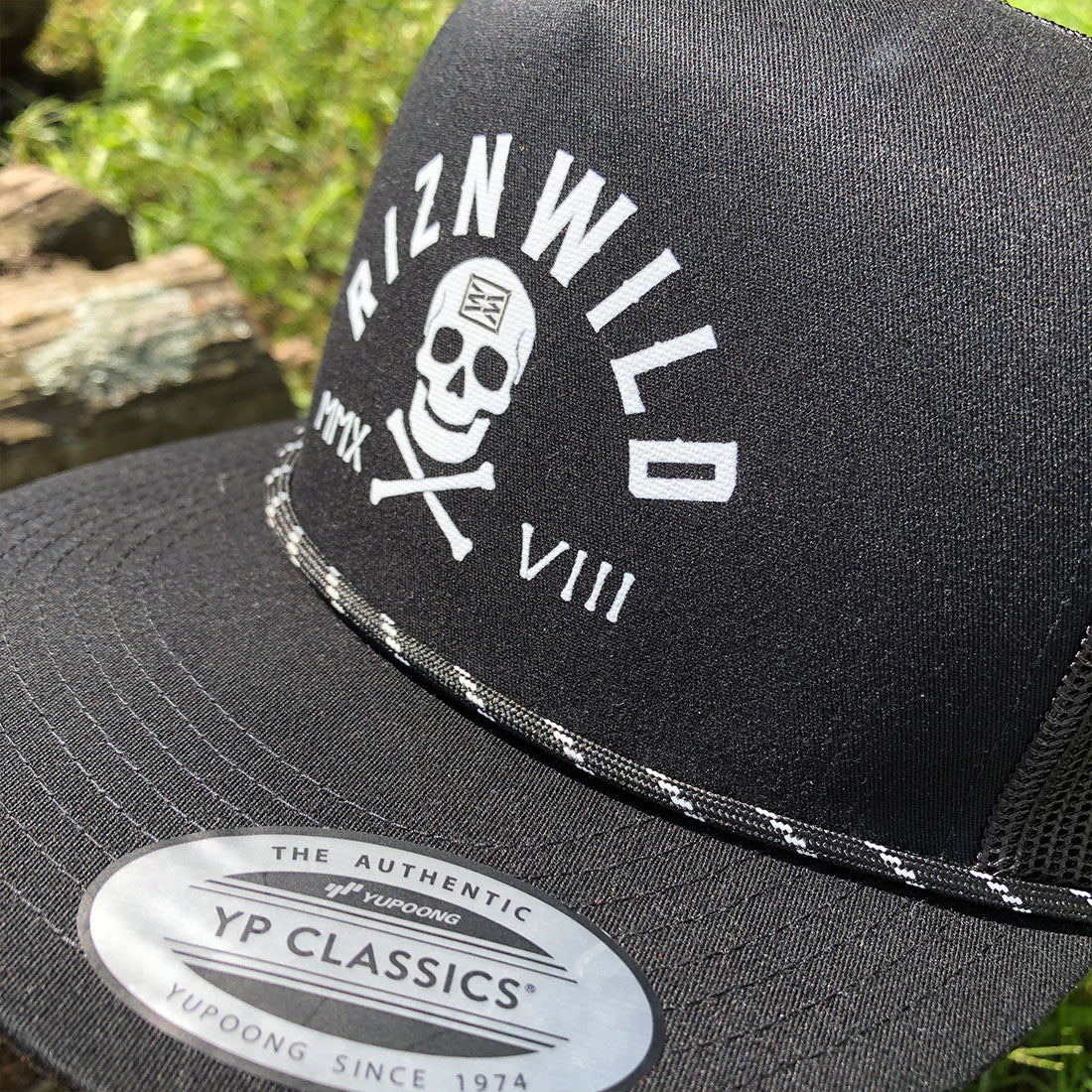 A close-up of a black trucker hat with a white embroidered skull and crossbones design on the front panel, featuring the text 'RIZNWILD' and 'VIII'. The brim has a visible white and black patterned trim, and a silver and black sticker for 'YP Classics by Yupoong' is attached.