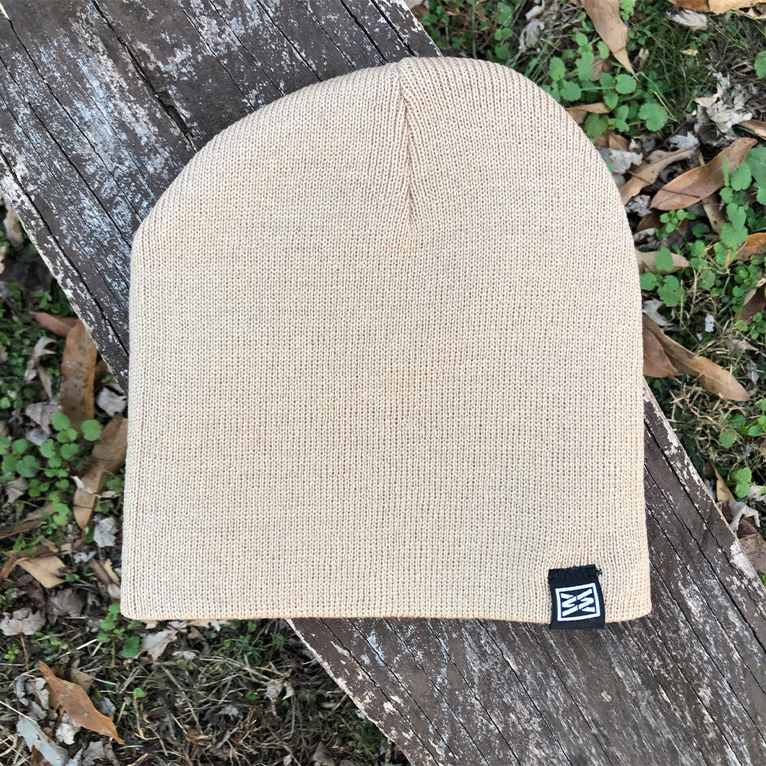 Beige ribbed knit beanie with logo tag on wooden surface outdoors